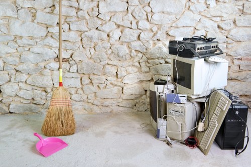 In a room with a rough, light-colored stone wall, there is a small pile of electronic waste including a computer monitor, a desktop tower, an old keyboard, and a television, all stacked together on the floor. Nearby, a pink plastic dustpan and a yellow and orange broom with natural straw bristles are positioned on the concrete floor, which has a neutral tone. The electronic equipment appears secondhand and disorganized, with cables visible draping over the devices. The setting suggests a temporary or storage area for rubbish awaiting collection or disposal, highlighting clutter typical of domestic or commercial waste removal carried out by Business Waste Removal Bethnal Green. The environment is well-lit, with natural light filling the space, emphasizing the textures of the stone wall and the various materials of the waste items. This scene reflects the kind of rubbish suitable for professional clearance services in Bethnal Green, focusing on electronic waste disposal and general clutter removal in an indoor setting within the postcode area.