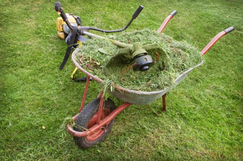 A red and black plastic garden wheelbarrow filled with freshly cut grass and plant debris, positioned on a patch of well-maintained green grass. The wheelbarrow's handles extend to the right, with one handle partially visible, while the curved metal support and wheel are at the front. A small, green grass trimmer with a black handlebar and a yellow attachment is resting on top of the grass in the wheelbarrow, with the trimmer's shaft extending upward and to the left. The surrounding environment appears to be an outdoor residential or garden area, characterized by lush, evenly cut grass. The scene suggests ongoing garden maintenance, with the presence of garden tools and waste materials, making it relevant to rubbish removal services in local areas such as Bethnal Green, London.