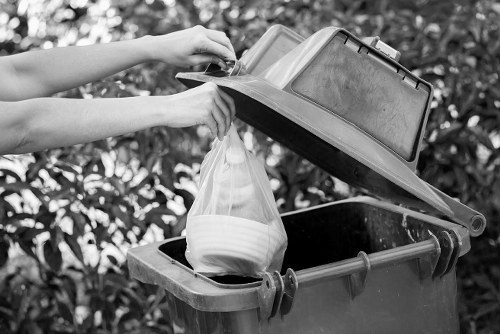 A person is seen lifting the lid of a grey waste bin situated outdoors on a paved area, with a background of dense leafy bushes. The individual is placing a transparent plastic bag filled with household waste into the bin, which appears to be made from durable plastic with a textured surface. The bin has a black interior and is equipped with a hinged lid, which is partially open and slightly tilted. The waste bag contains various discarded items such as white paper or cardboard bowls and other light-colored refuse, suggesting general household rubbish. The scene is well-lit by natural daylight, with shadows cast on the bin and the person's arms, indicating an outdoor environment. This setting reflects typical rubbish collection contexts in Bethnal Green, supporting local waste management services that handle general rubbish disposal for businesses and residents in the area.