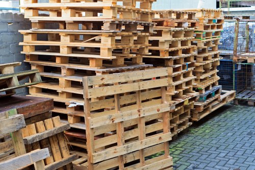 A large stack of untreated wooden pallets arranged outdoors on a paved surface near a building, with some pallets leaning against a wall on the left side. The pallets vary in size but primarily consist of smooth, light-colored timber with visible grain and occasional darker knots. The pallets are stacked in multiple layers, creating a tall, uniform pile occupying most of the right side of the image. In the background, a metal fence partially encloses the area, and there are glimpses of other industrial or storage structures. The natural light suggests daytime, with shadows cast on the ground, emphasizing the textures and arrangement of the pallets. This scene reflects a typical outdoor setting for waste disposal or storage of timber for recycling or reuse, relevant to businesses involved in rubbish removal services in Bethnal Green or surrounding London areas. The overall impression is of a well-organized pallet storage site suitable for waste management or logistics operations.