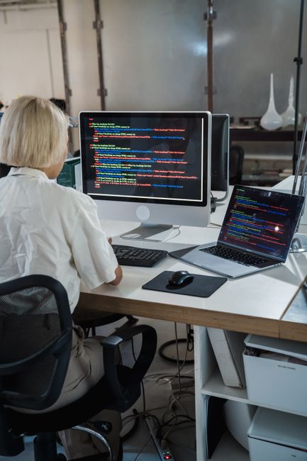 A close-up photograph of a computer monitor displaying lines of code in a dark-themed IDE, featuring syntax highlighting with various colors such as orange, blue, and white. The code includes HTML and