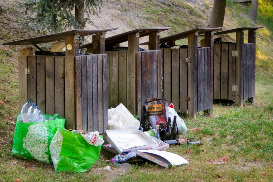 A collection of overflowing rubbish bins and scattered waste on a paved urban sidewalk, with various black, black, red, and grey bins filled with mixed waste, including paper, cardboard, plastic, and